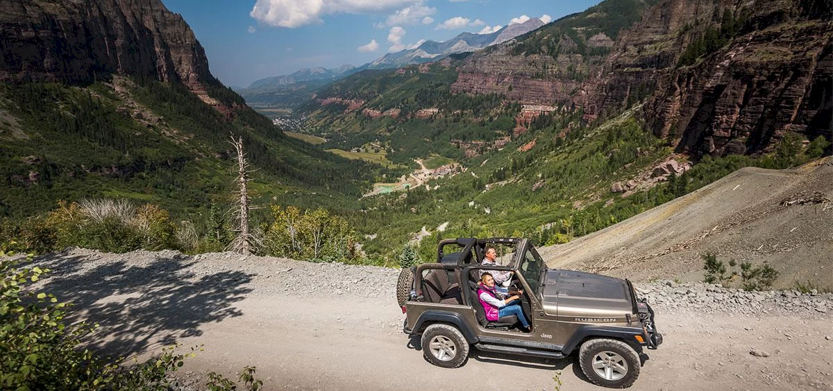Two people in a Jeep with the top off drive along the cliff of Black Bear Pass.