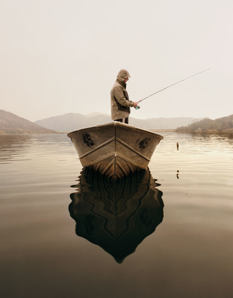 Man stands in small boat on lake with fishing rod in hand.