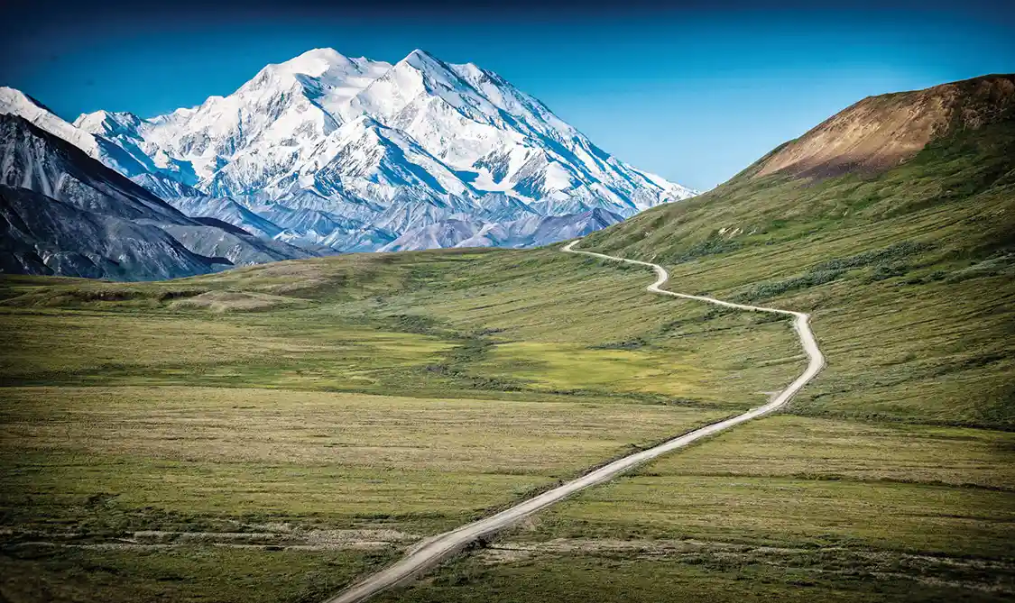 Winding dirt road runs toward a snow covered white mountain, called the Denali Highway.