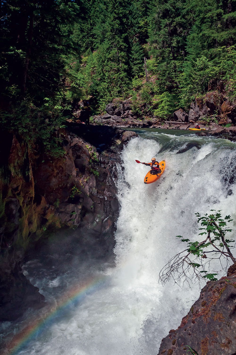 kayaker heading toward a rainbow at the bottom of a waterfall