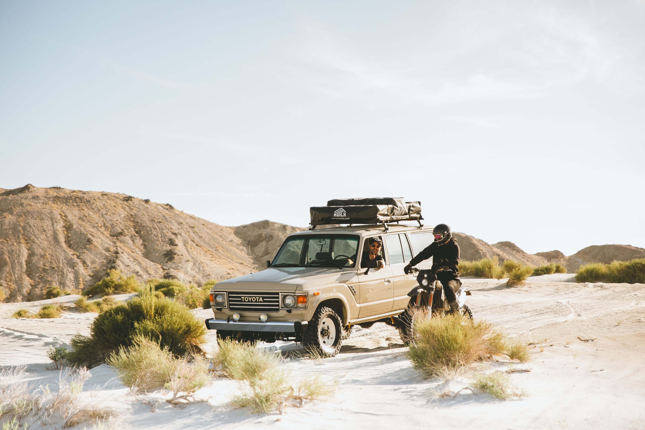 Man in Land Cruiser and motorcycle rider talk while parked in sand.