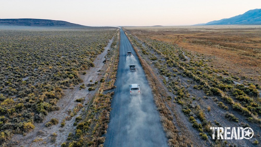 Three adventure vehicles head out to the lands less explored via dirt roads and sagebrush on both sides. Far off hills greet them. 