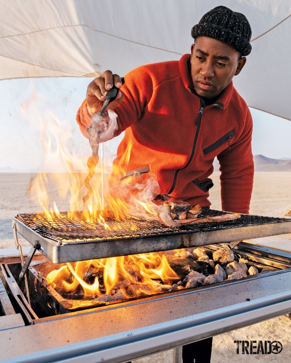 Frazier, earing an orange fleece from Camp Yoshi, cooks bacon over an elevated fire grill.