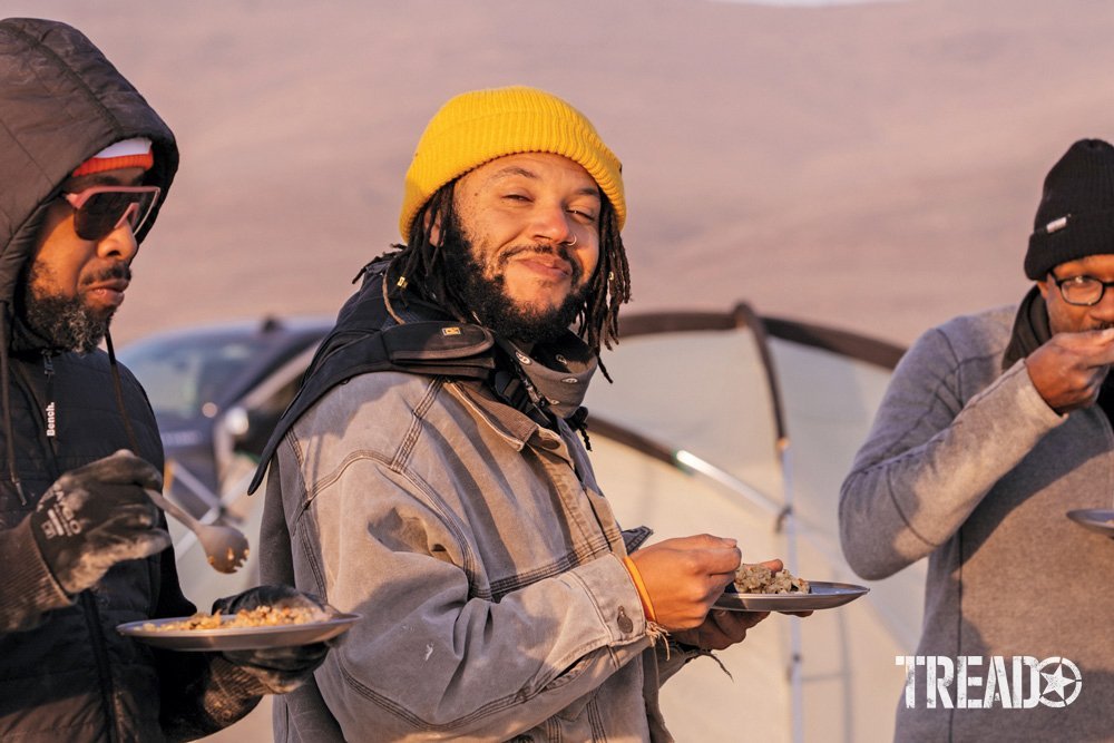 Three Camp Yoshi attendees are enjoying eating dinner, as the hold their plates in hand, standing in front of tents.