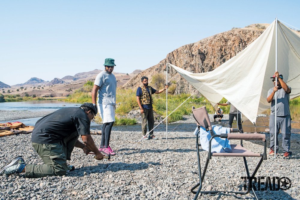 Two men hold up a white awning while another secures its guy lines into the rocky ground.