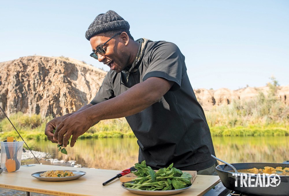 Rashad Frazier, wearing a hat and t-shirt, smiles as he dresses his camp meal with herbs.