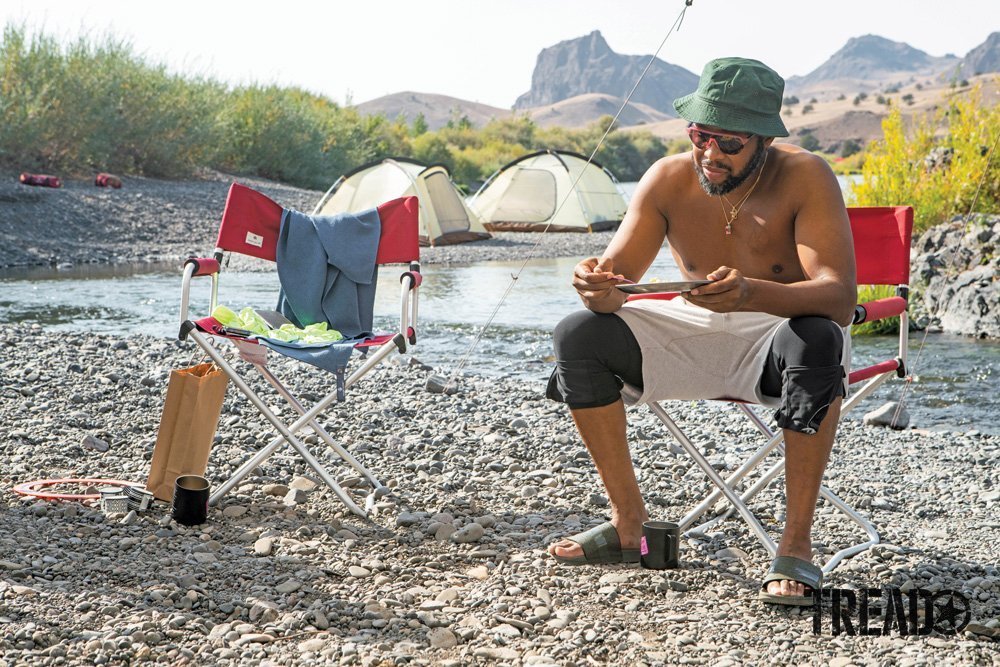 A Camp Yoshi participant enjoys camp food next to a stream while sitting in a red camp chair with tents set up in the distance.