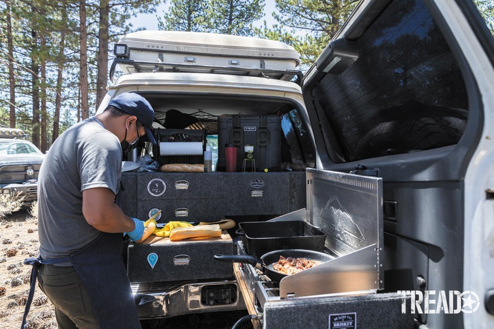 Man cooks using camp kitchen setup in back of vehicle.