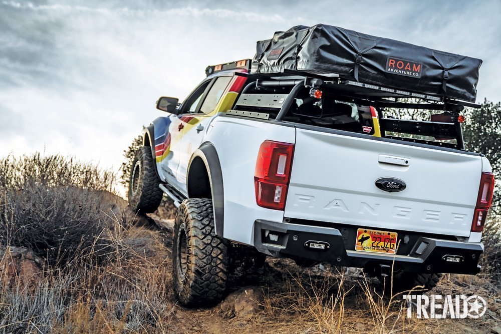 The multi-colored customized Ford Ranger showcases a truck bed rack, roof top tent, and is climbing a hill. 