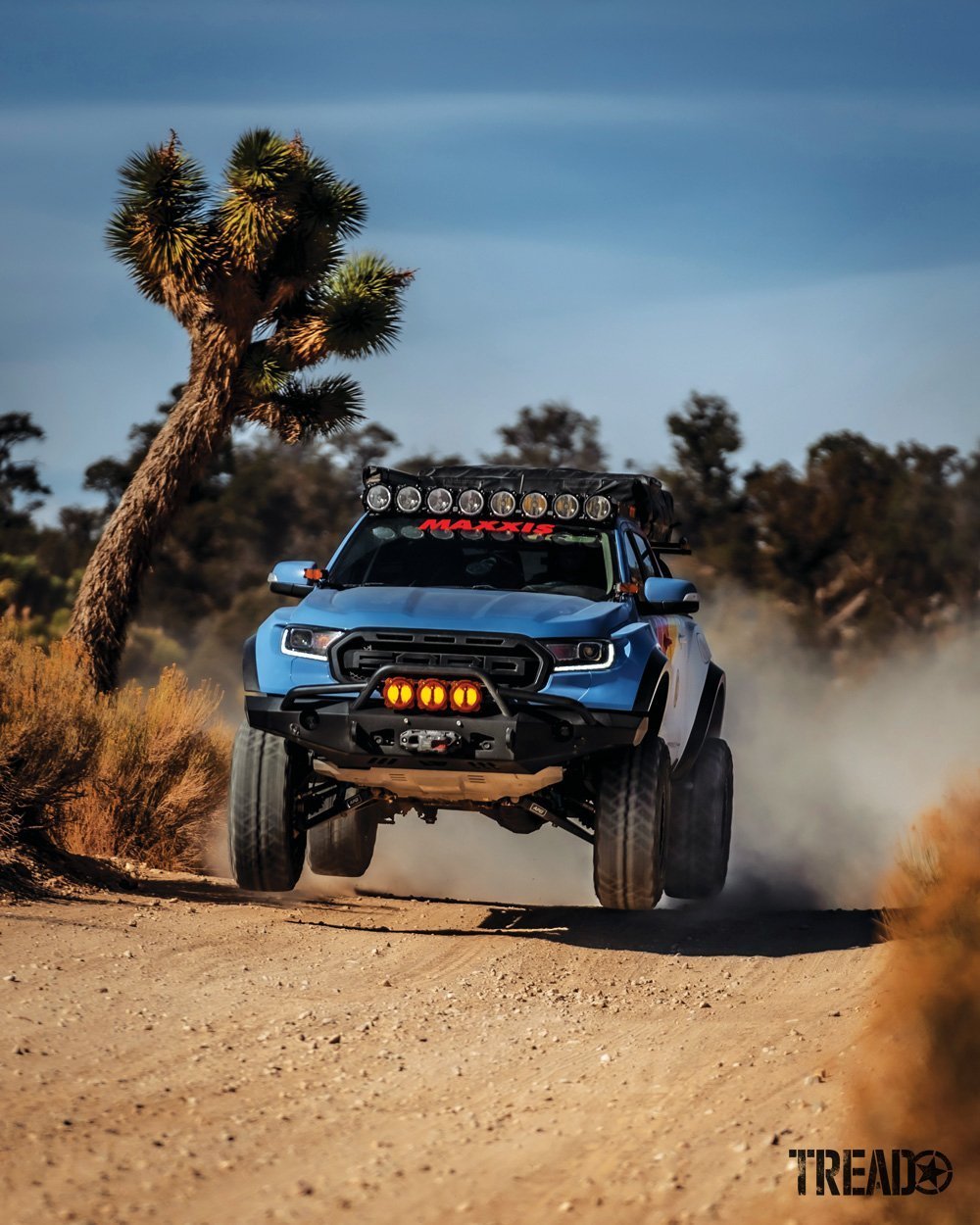 2019 Ford Ranger action shot as it drives down a desert dirt road, its light blue front end accented by yellow aux lights. 