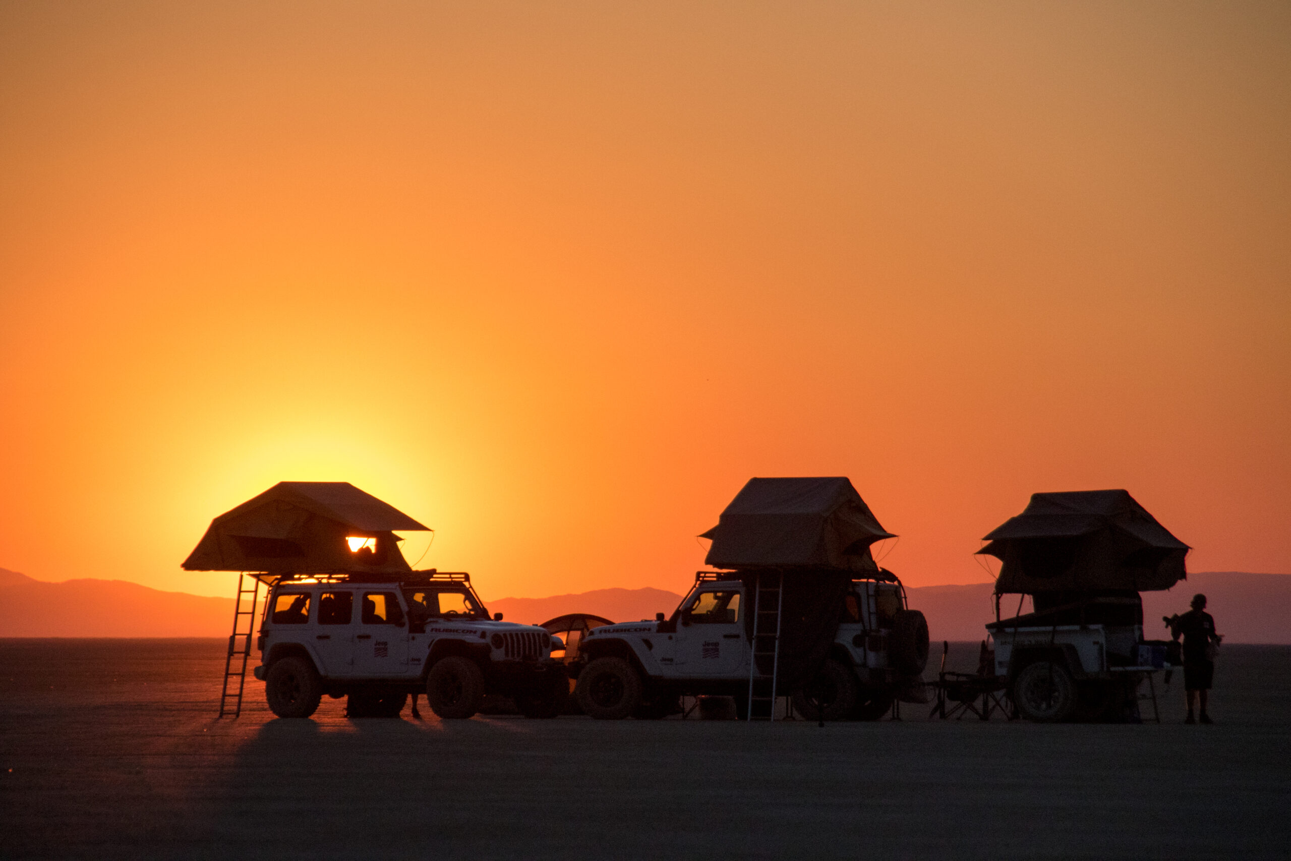 Two jeeps with rooftop tents camp on a dry lake bed at sunset.