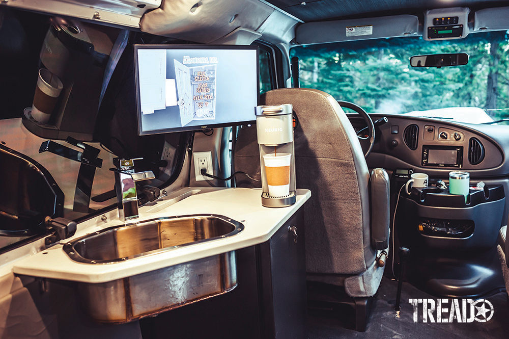 A Keurig coffee maker, as well as a sink and monitor on a white countertop, is anchored next to the van's window on the interior.