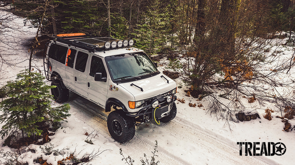 Two solar panels and orange recovery boards are mounted to a platform roof rack on a white 4x4 van.