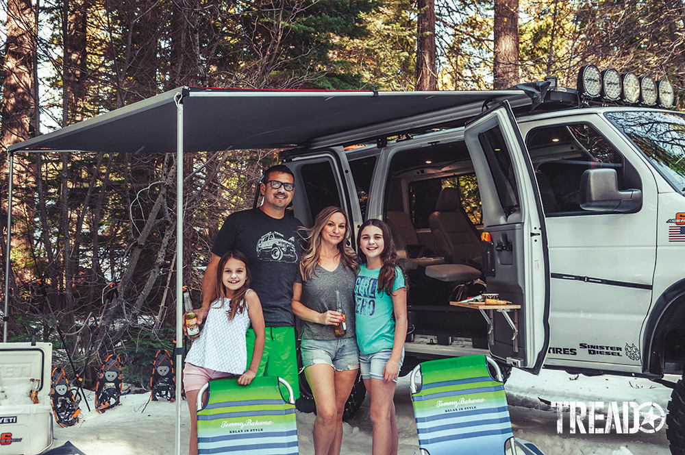 The white Ford E350 Super Duty 4x4 van sits behind Ken Montes and family, leaning on striped camp chairs.