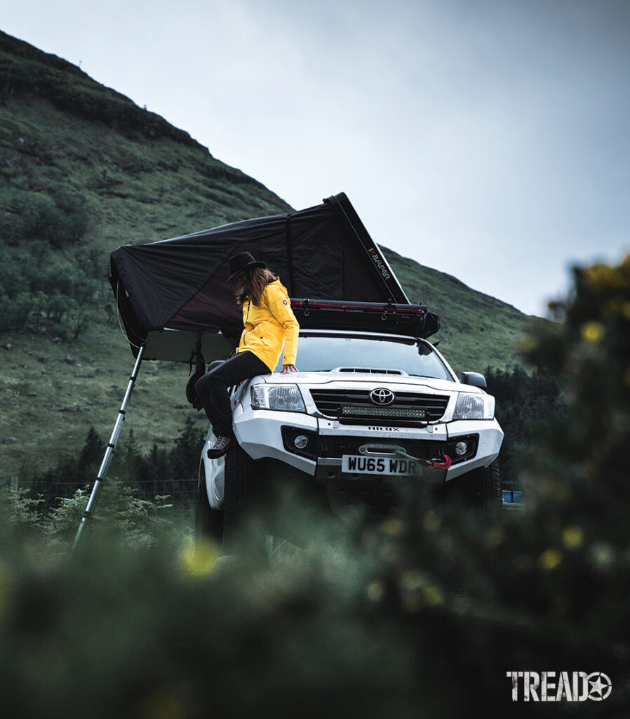 Woman sitting on customized white Toyota with rooftop tent open. She is wearing a hat, yellow jacket, and black pants.