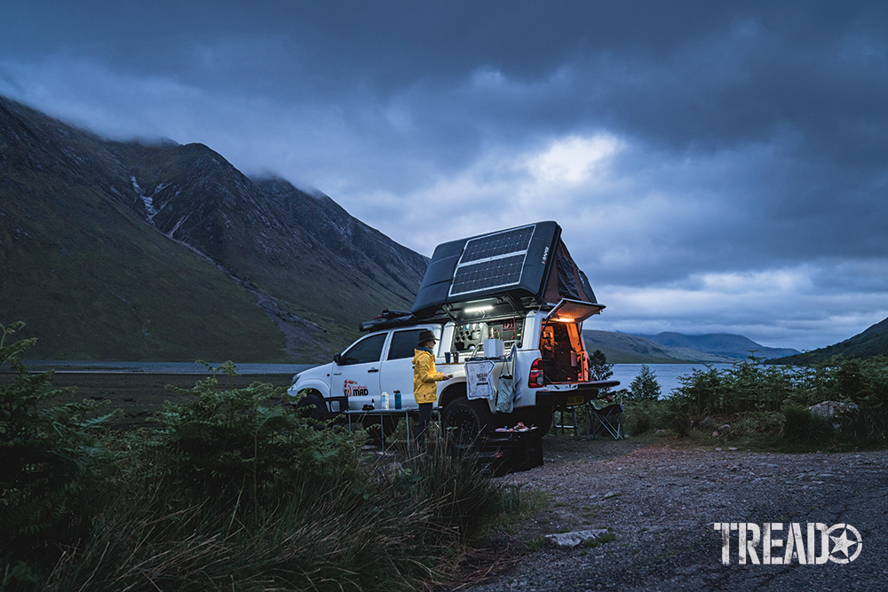 Preparing dinner next to a customized white Toyota and rooftop tent while overlooking the water and nearby green hills.