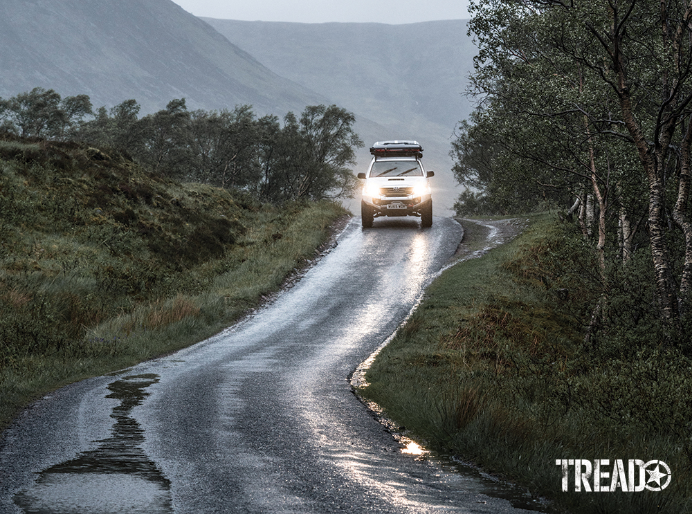 Misty , raindrops shone like jewels on the windscreen of a customized white vehicle as it drives down a paved road with hills in the background.