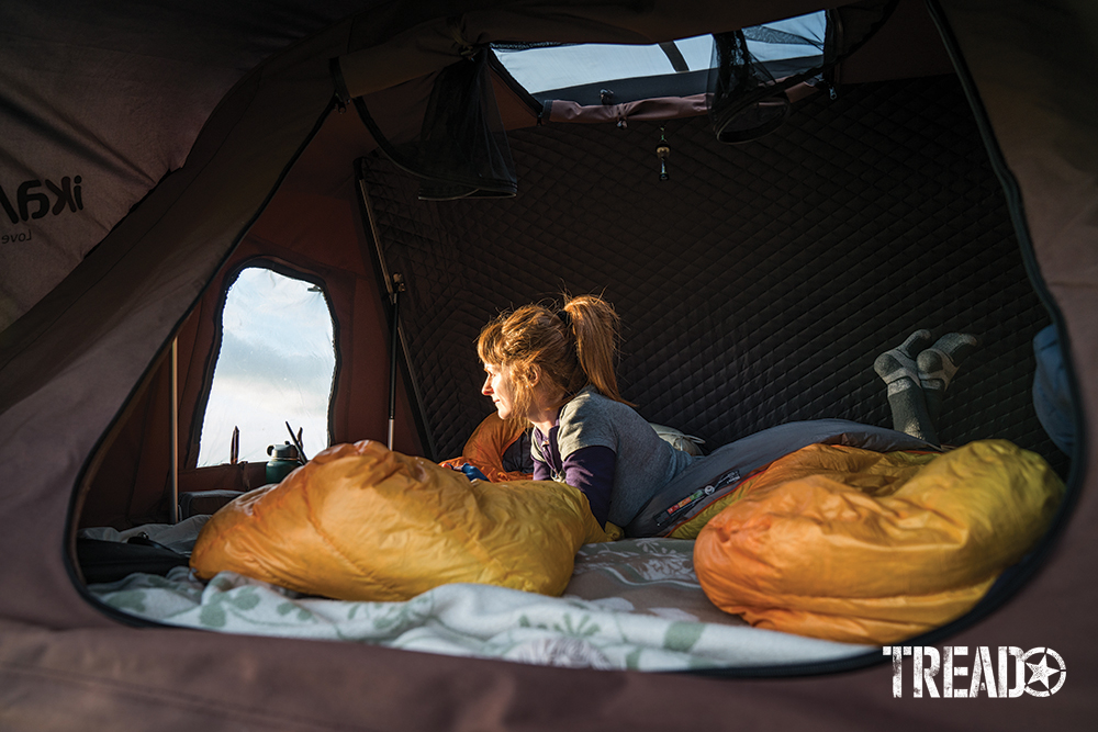 Opened rooftop tent with woman looking outward, two yellow sleeping bags laid out ready for sleep.