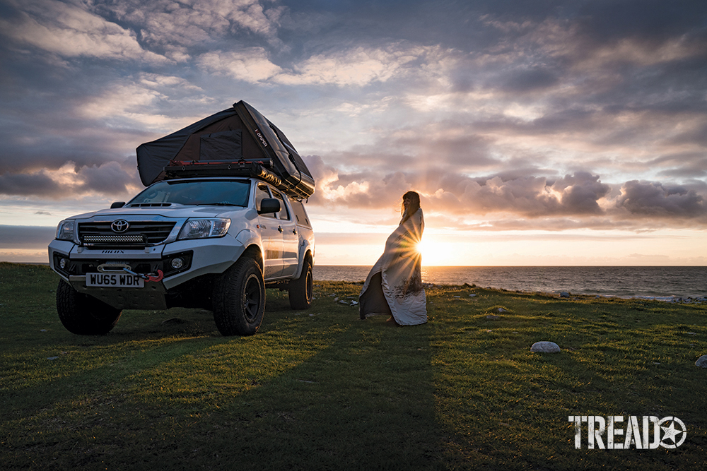 A white customized Toyota with rooftop tent sits on green grass next to water with the sun setting in clounds and behind woman in blanket.