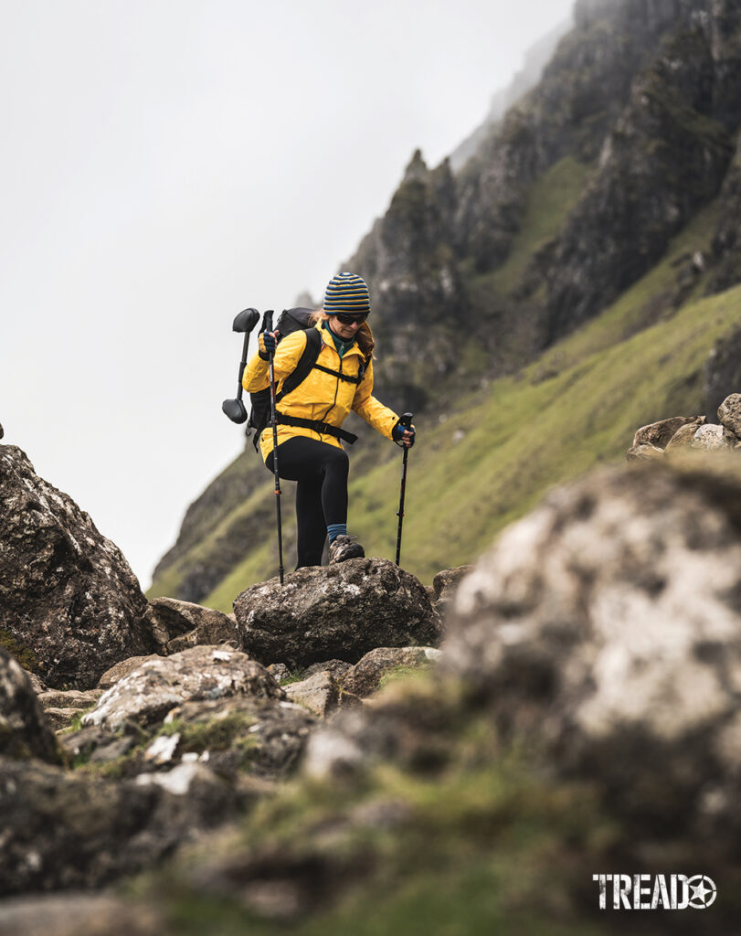 Hiker in yellow jacket, black pants, and backpack hikes a Scottish landslide area on big rocks.