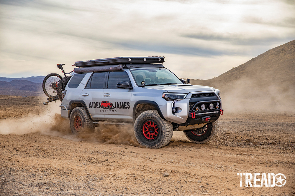 A silver customized 2017 Toyota 4Runner with slim rooftop tent, red wheels, drives through dusty conditions.