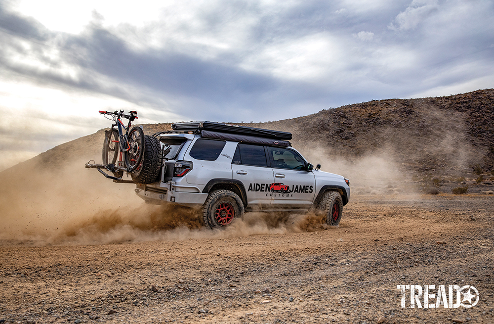 A silver customized 2017 Toyota 4Runner with slim rooftop tent and red wheels, slashes through the dirt.