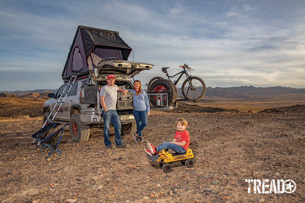 Customized 2017 Toyota 4Runner has rooftop tent opened and tailgate opened while the Fisher family stands in front of it with young boy sitting on bulldozer toy.