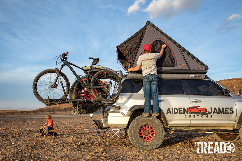 A child with bulldozer toy plays next to a silver adventure 4Runner with bicycles and man zipping up rooftop tent window.