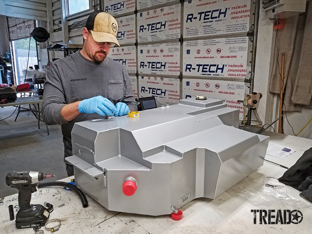 The owner of Wanderlust Overland, wearing a baseball cap and gray shirt, preps the Delica Space Gear replacement tank before installation.