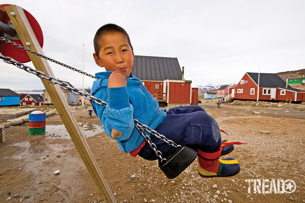 Inuit boy swinging on swing set wearing clue jacket and colorful boots smirking at camera.