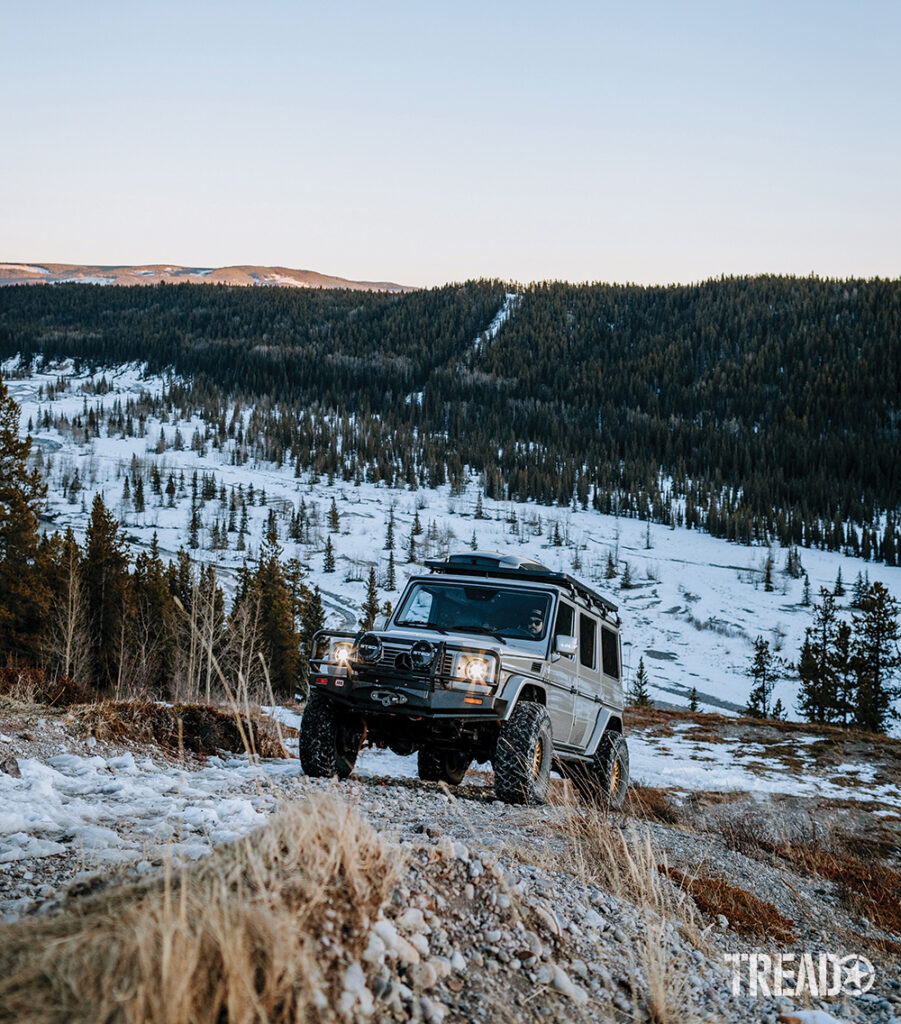 The silver Mercedes G-Wagen heads uphill on a partially snow-covered trail with hillside of pine trees behind it.