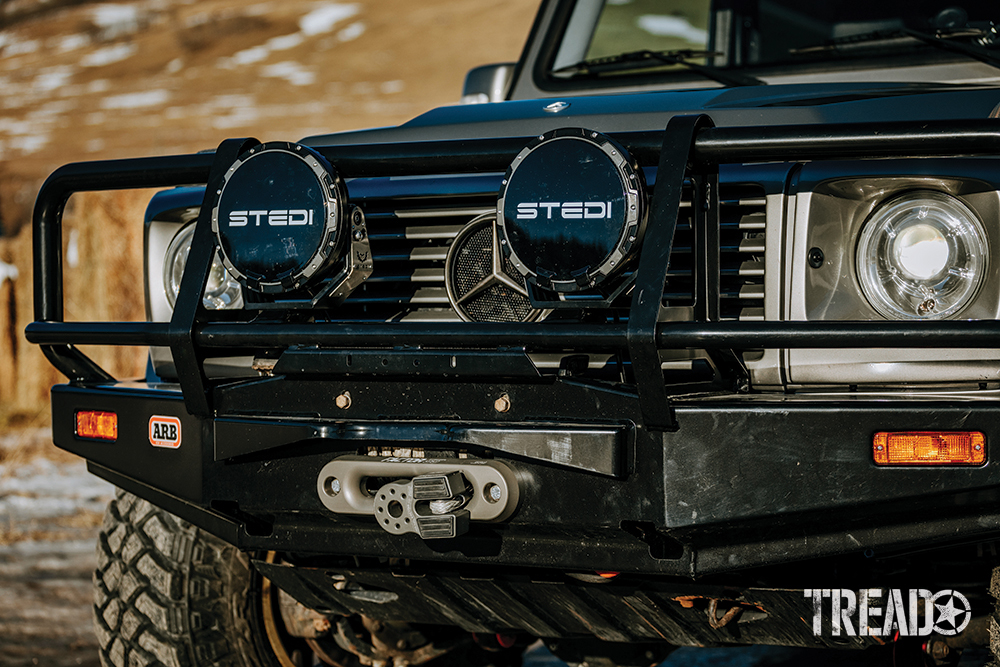 Front winch bumper, bull bar, and black-covered aux lights on silver Mercedes G-Wagen.