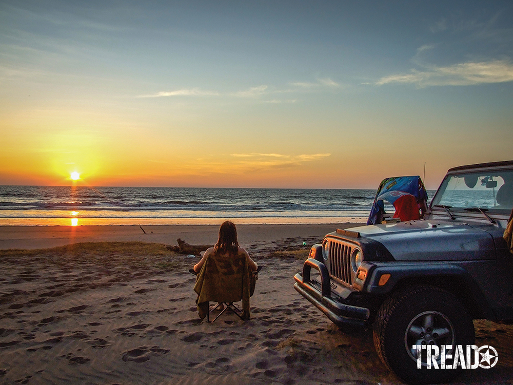 Man sitting in chair on beach watching sunset while silver Jeep is next to him.