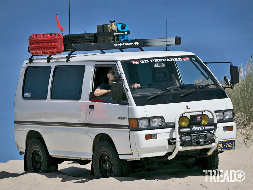 A white third-gen Mitsubishi Delica is parked in the sand. Driver sits on the right side of this right-hand drive vehicle.