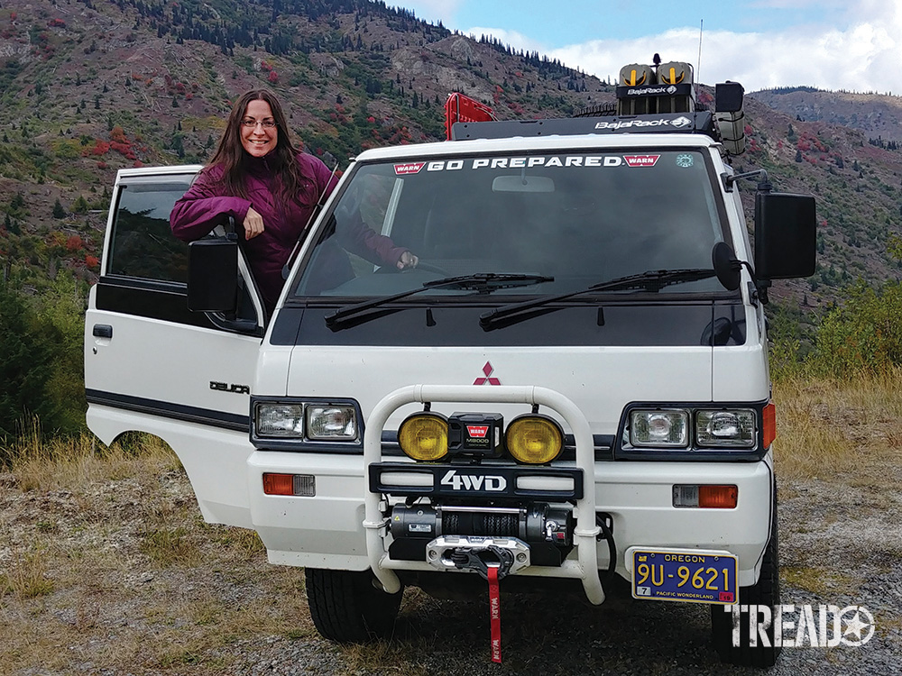 Driver hangs out the driver side door of a right-hand drive white Mitsubishi Delica van.