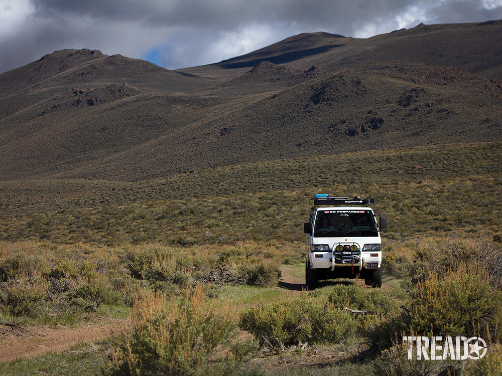 A white Misubishi Delica van is parked along a dirt road with mountains in the background.