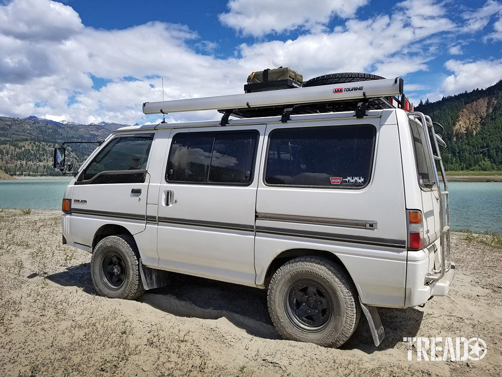 A white Mitsubishi Delica Star Wagon is parked in the sand next to a river..