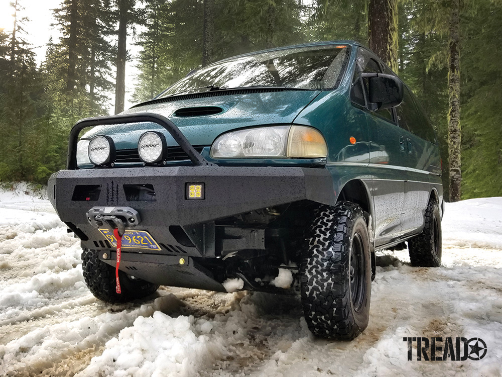 The front end of a green Mitsubishi Delica van with an off-road front bumper, lights, and winch.