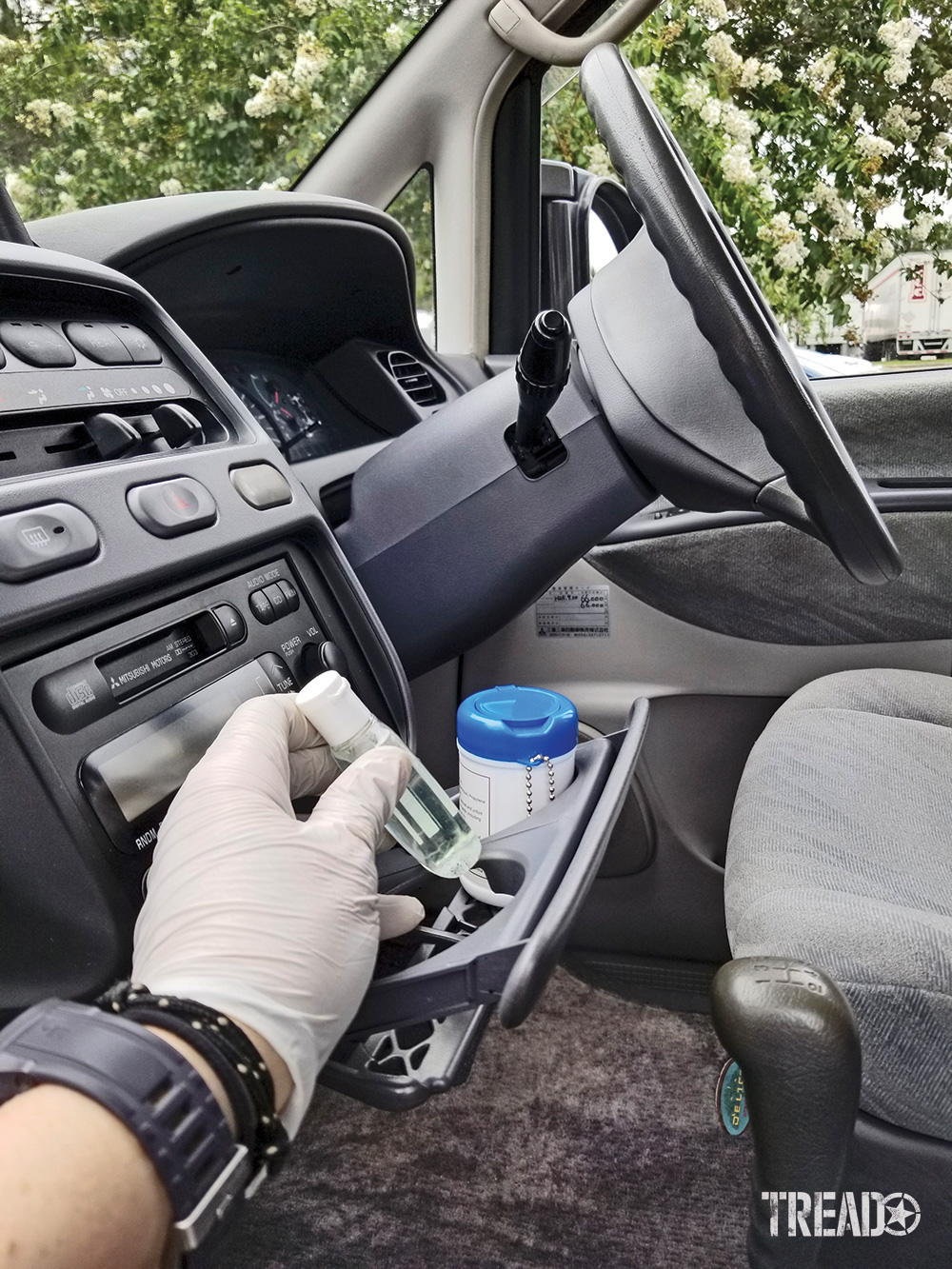 The front seat of a right-hand drive Mitsubishi Delica L400 is seen with a gloved hand grabbing hand sanitizer from the pull out cup holder.