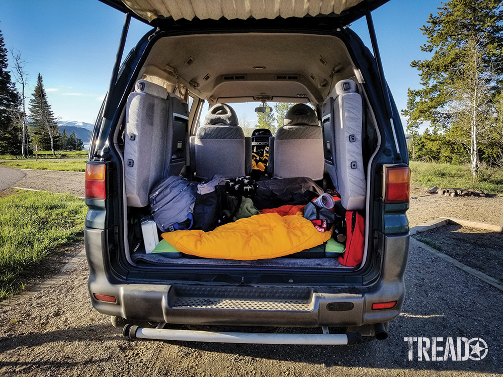 The inside view of the Mitsubishi Delica L400 with third row seats folded up towards the side and mid-row seats folded up towards the front. Sleeping gear is in the back to show how users can sleep on the floor.