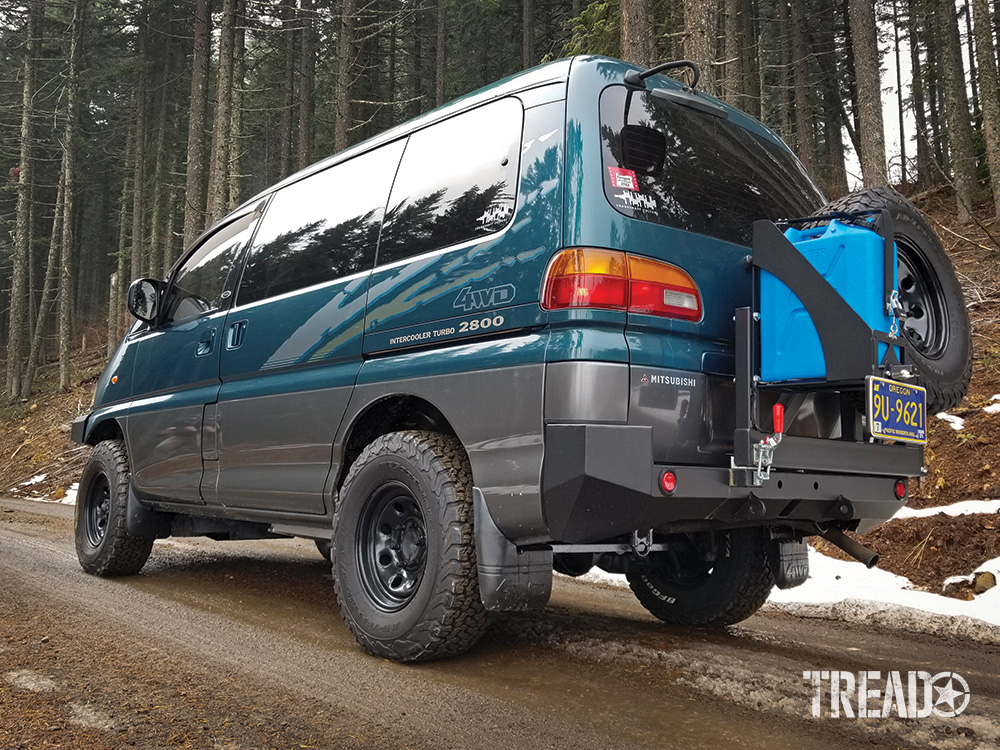 A rear quarter view from the driver side of a green Mitsubishi Delica van with a swing out bumper that holds the spare tire and water can.