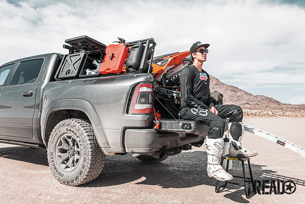 Man sits on tailgate of truck with dirt bike in back and bed rack storage holding gear shown.