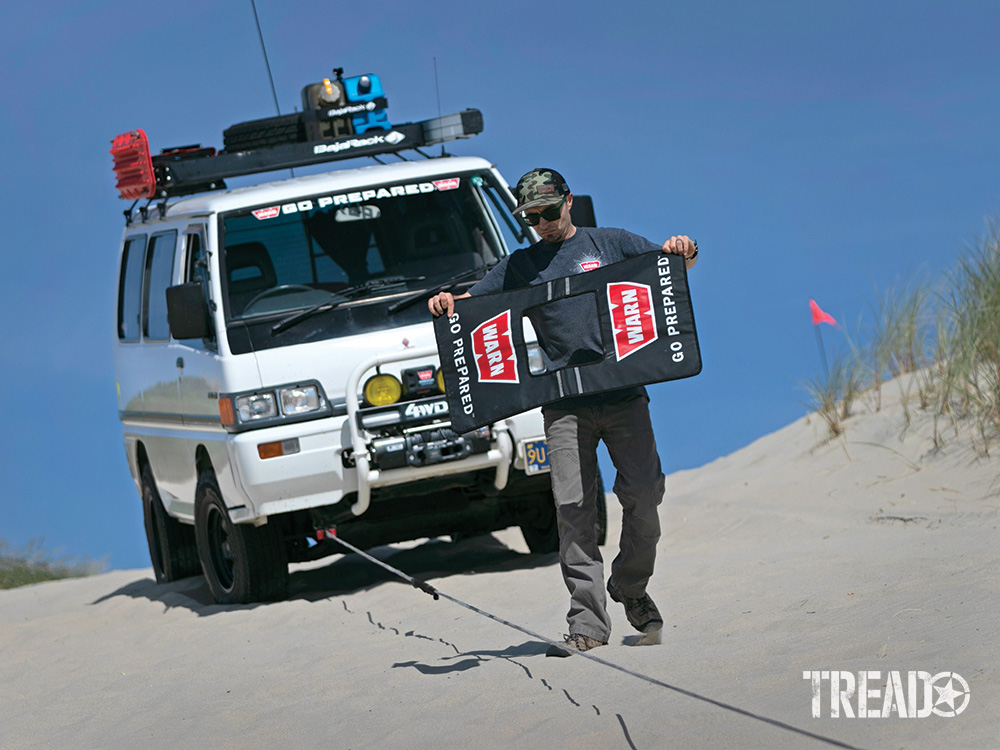 A man uses the winch on a white Mitsubishi Delica that is parked in sand with it's winch lead extended.