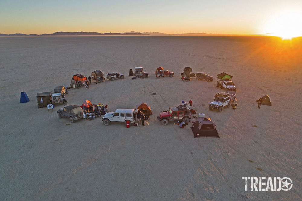 A group of customized 4x4s collect in a circle on a dried lakebed as the sun sets.