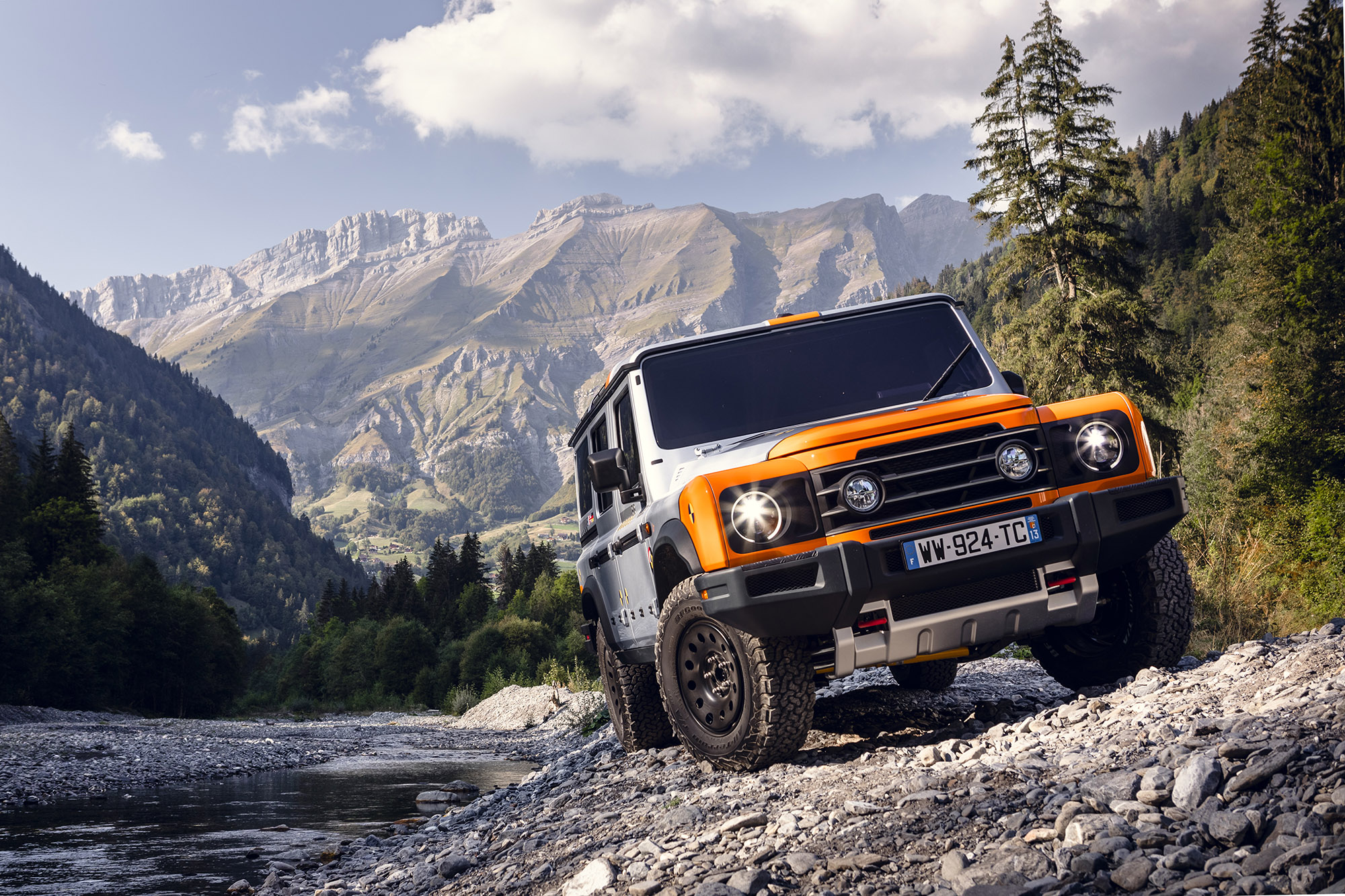 An orange and silver INEOS Grenadier prototype traverses a rocky mountain trail with mountain range behind it.