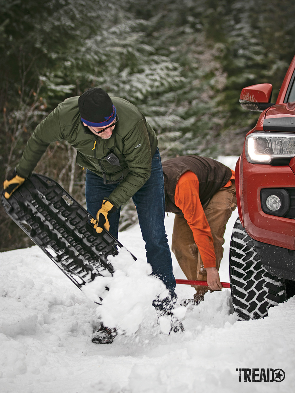 Two men use a black recovery board and red shovel to dig out an orange Toyota truck in deep snow.