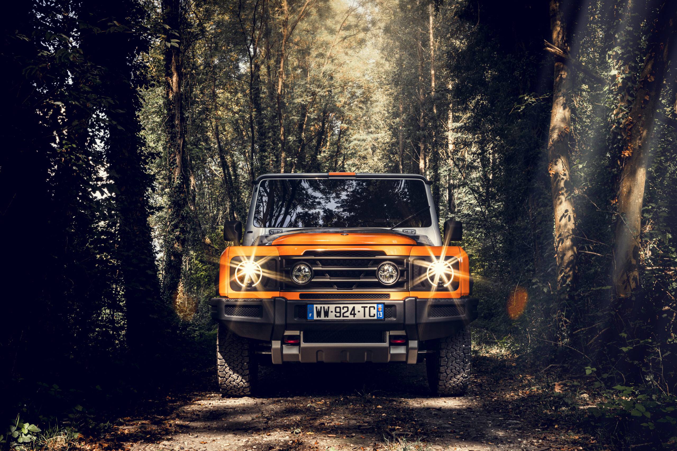 The orange and silver INEOS Grenadier's front end with lights on in a thick forest.