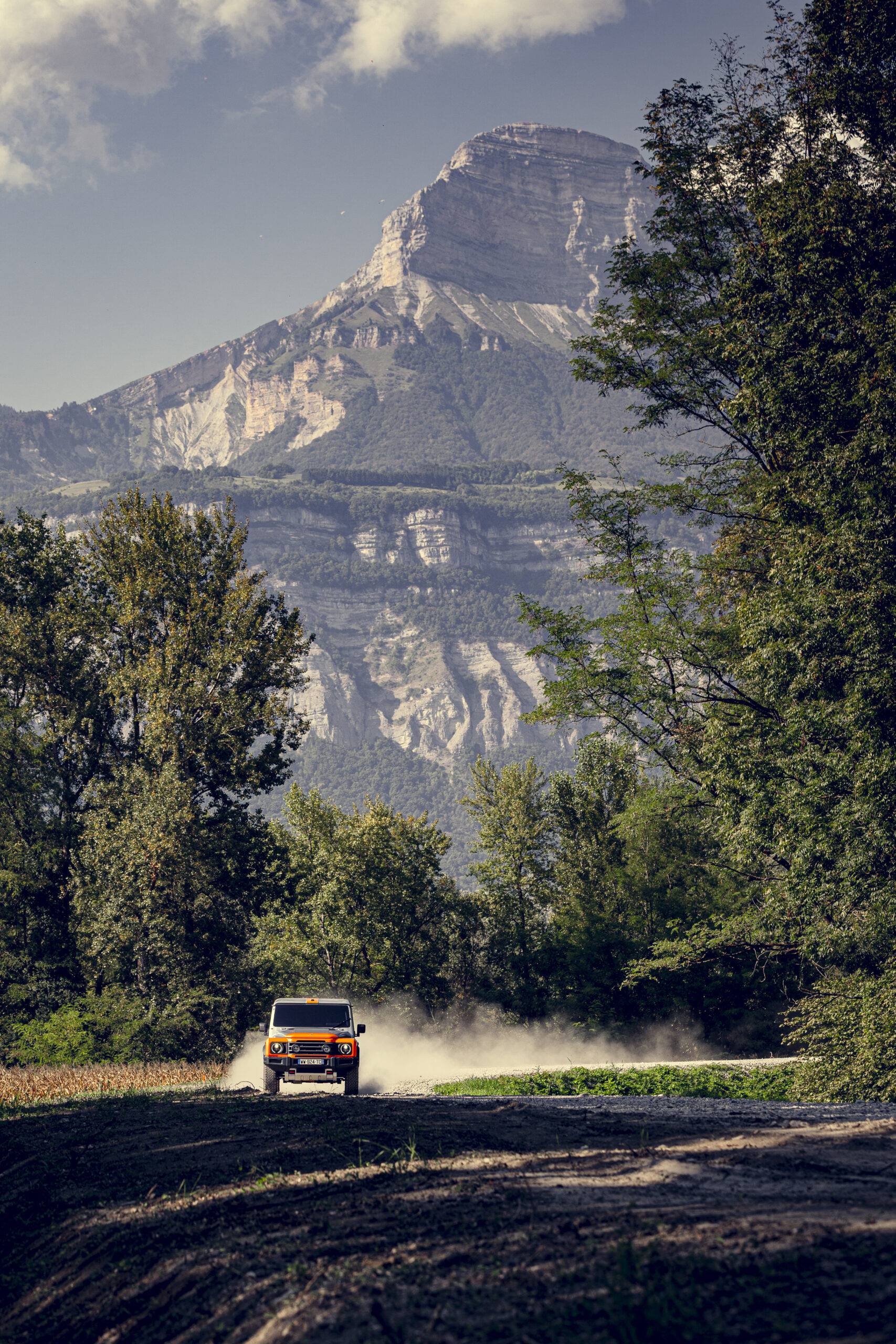 The orange and silver INEOS Grenadier drives on dirt road in front of mountains