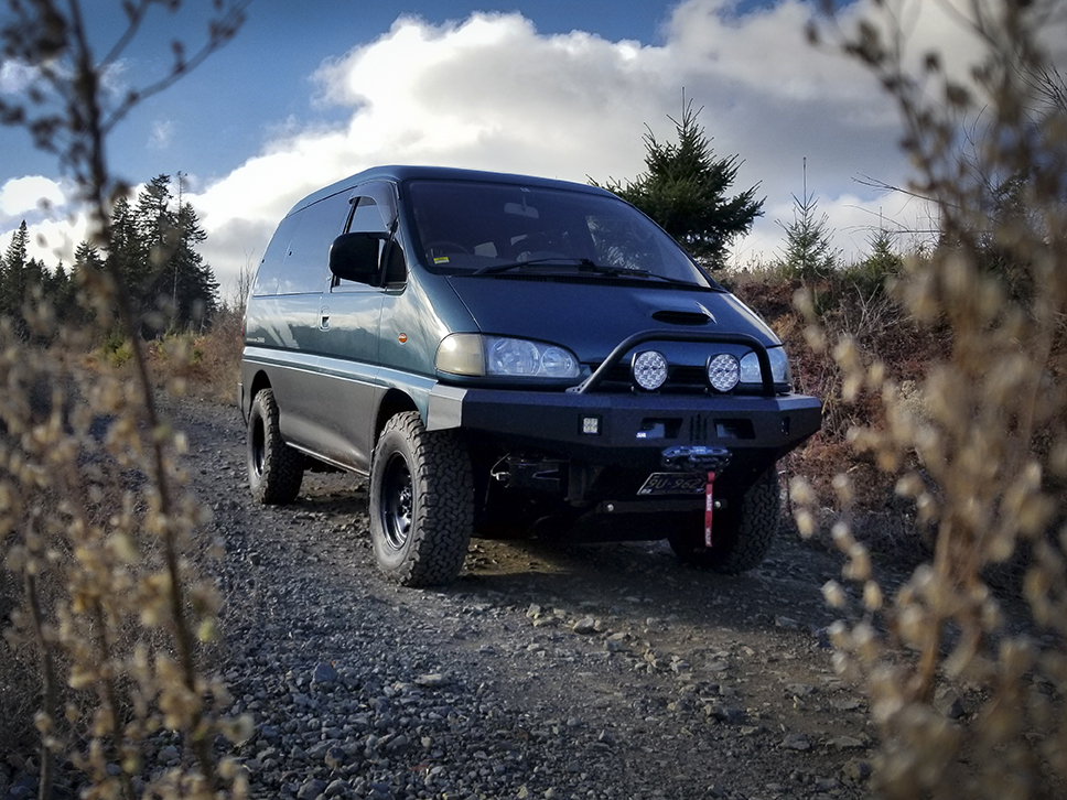 Green Mitsubishi Delica van parked on dirt with bushes in forground.