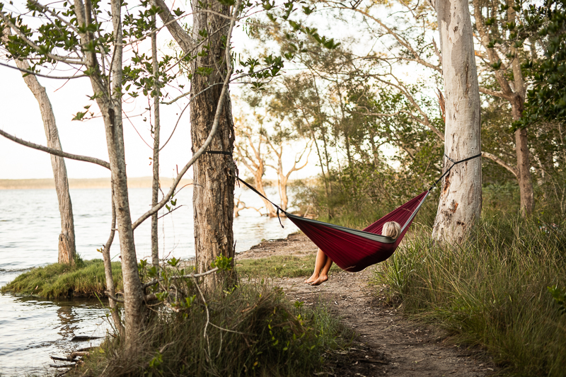 person lays in red hammock tied between two trees next to a lake